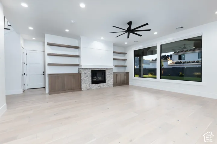 Living area featuring light-toned wood-finish flooring, a stone-surround fireplace, built-in lower cabinetry with floating shelves, large picture windows, and a multi-blade ceiling fan