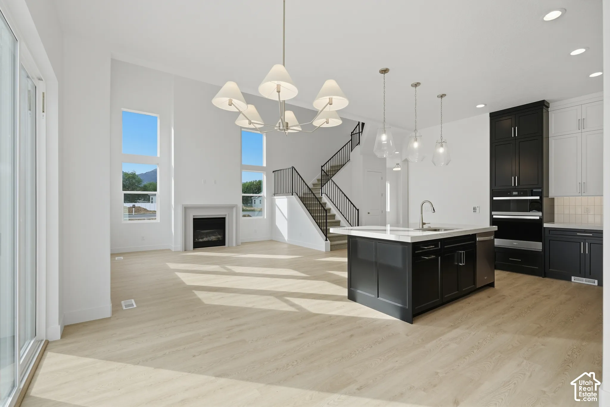 Open-concept interior featuring light wood-finish flooring, two-story windows, a fireplace with a minimalist mantel, a center kitchen island with quartz countertops, and a staircase with metal railings
