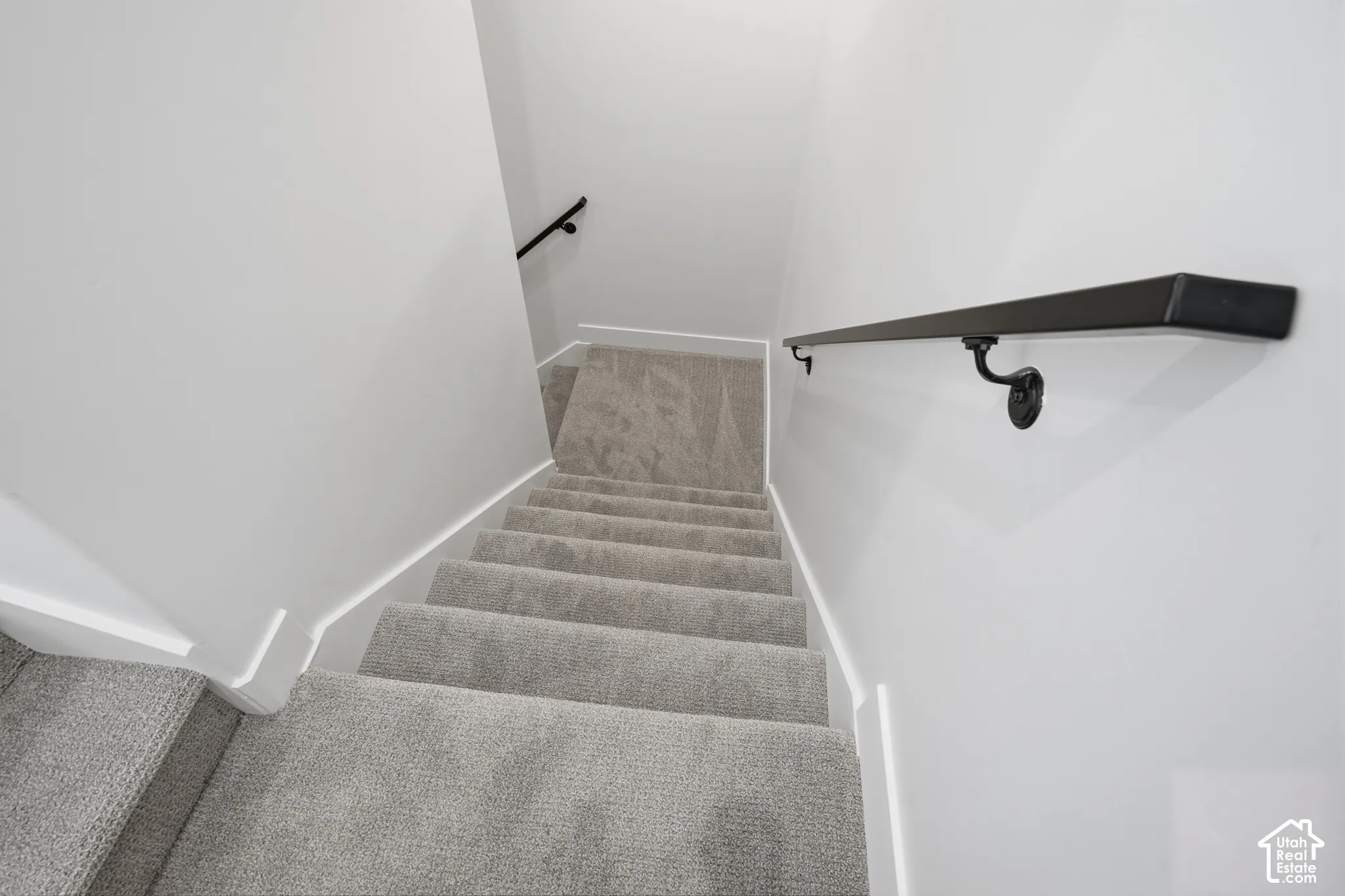 Staircase featuring neutral-toned carpeting, matte black wall-mounted handrails, and clean white walls