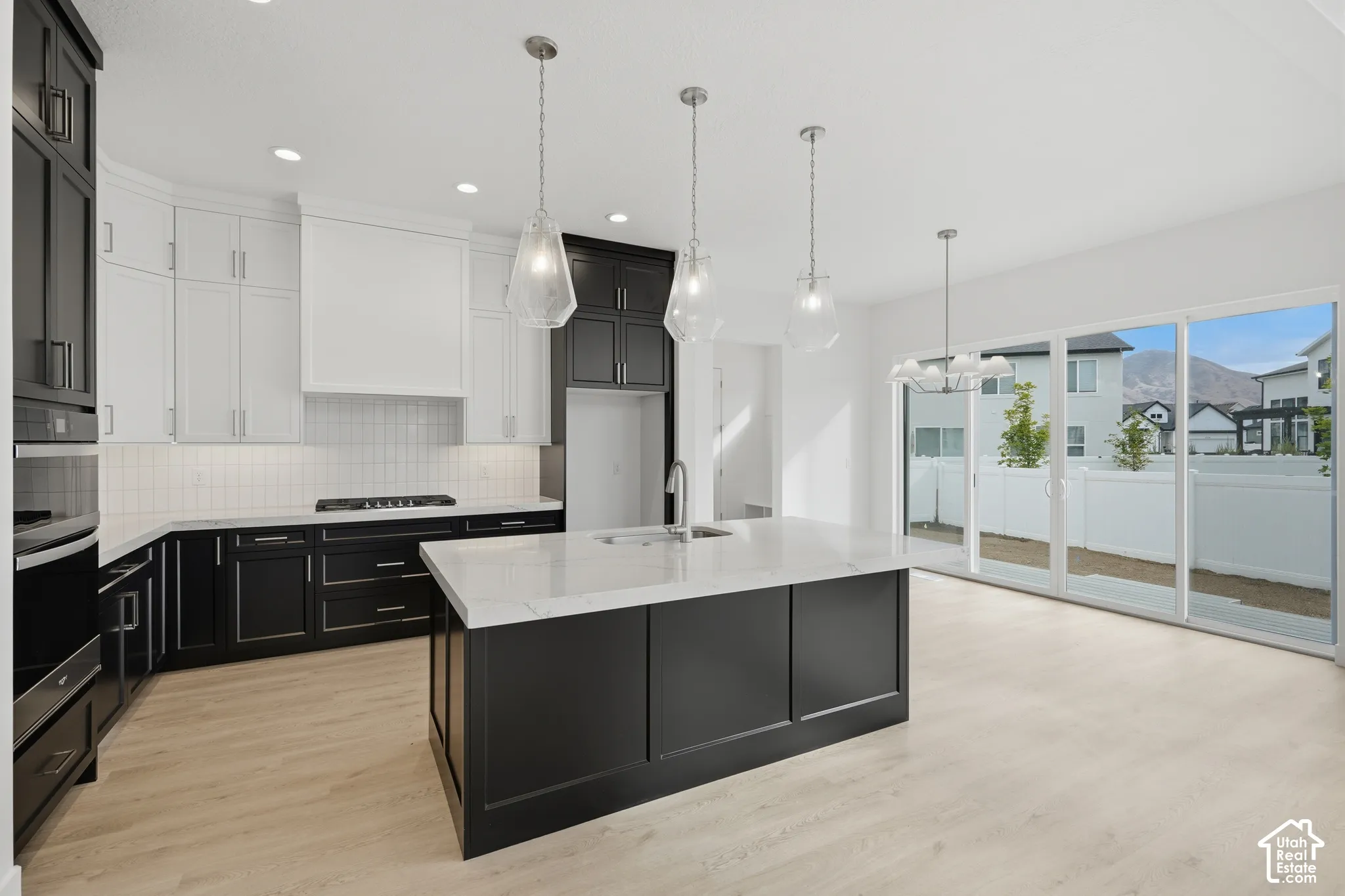 Two-tone cabinetry with contrasting white upper and dark lower units, expansive central island with quartz countertop, light-toned wood-finish flooring, recessed lighting paired with glass pendant fixtures, and large sliding glass doors