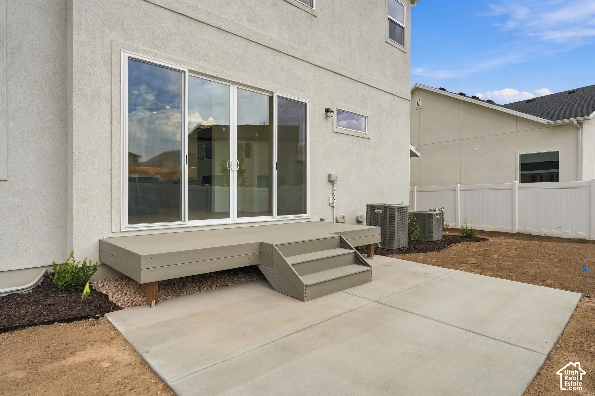 Stucco exterior featuring large sliding glass doors, a raised wooden deck with integrated steps, a poured concrete patio, and white vinyl privacy fencing