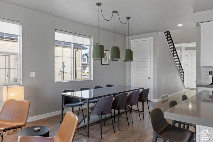 Open-concept interior featuring wood-finish flooring, trio of olive-toned pendant lights, large double-pane windows, and a staircase with a black metal railing