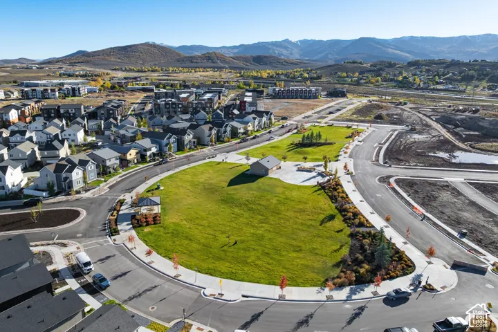 Aerial perspective of a community green space featuring manicured lawns, mature landscaping, paved pathways, a central pavilion structure, and mountain backdrops