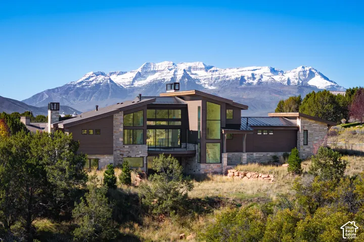 Contemporary architectural design featuring multi-level shed roofing, stone masonry exterior, expansive floor-to-ceiling windows, metal roofing accents, and a prominent mountain backdrop