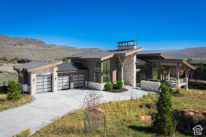 Modern mountain-style exterior featuring stacked stone masonry, a standing seam metal roof, multi-pane glass garage doors, timber support beams, and large-scale architectural windows
