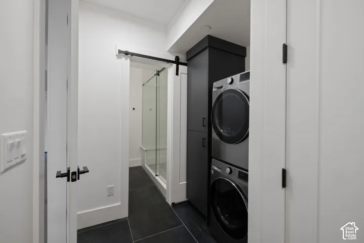 Stacked laundry appliances, tall charcoal-finished built-in cabinetry, dark tile flooring, and a sliding barn door entry