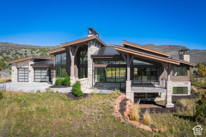 Modern mountain architectural design featuring natural stone accents, timber frame structural supports, expansive floor-to-ceiling windows, metal roofing, and an elevated deck with metal railing