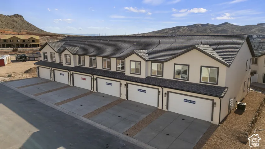Multi-unit residential exterior featuring a light-colored stucco facade, dark-toned concrete tile roof, gabled rooflines, and private multi-car garage bays, set against a backdrop of desert mountains