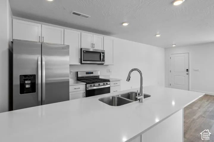 Kitchen featuring white shaker cabinetry, stone-finish countertops, stainless steel appliances, a central island with a double-basin sink, and wood-finish flooring
