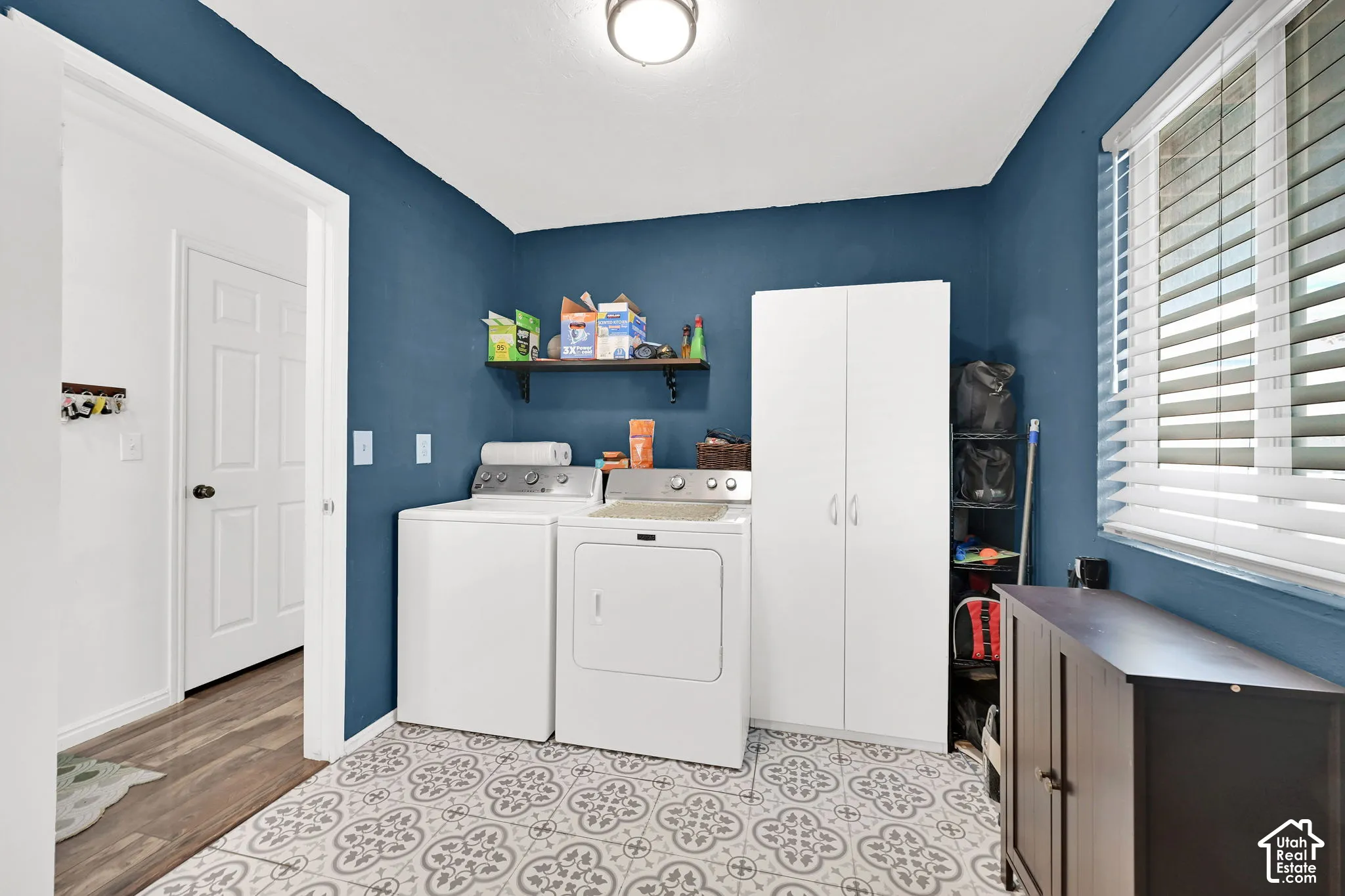 Utility room featuring patterned tile flooring, white built-in storage cabinetry, a wall-mounted wooden shelf, and a horizontal window with adjustable blinds