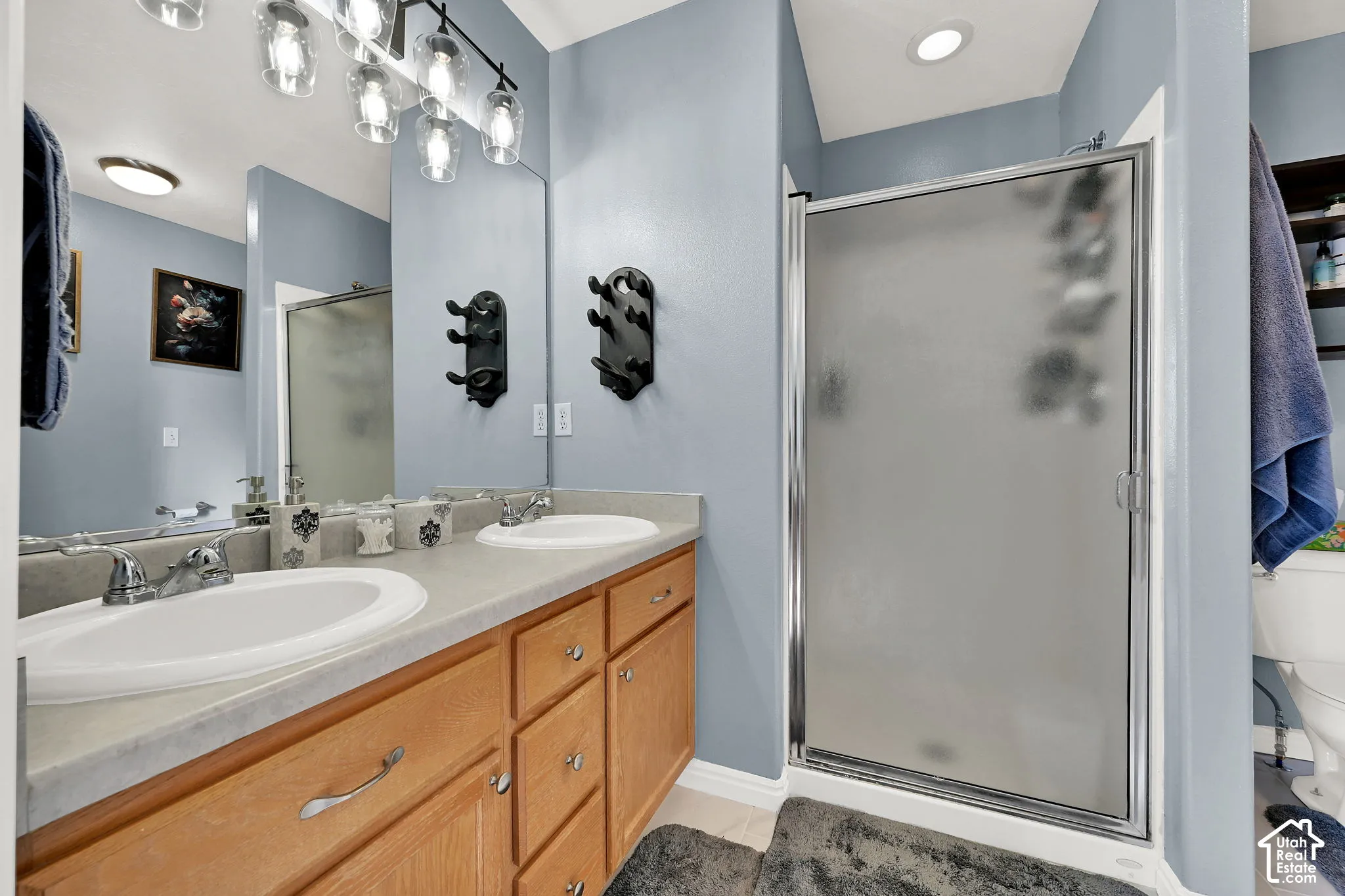 Bathroom featuring a dual-sink vanity with a solid-surface countertop, wood cabinetry, a walk-in shower with a frosted glass door, and multi-light pendant vanity lighting