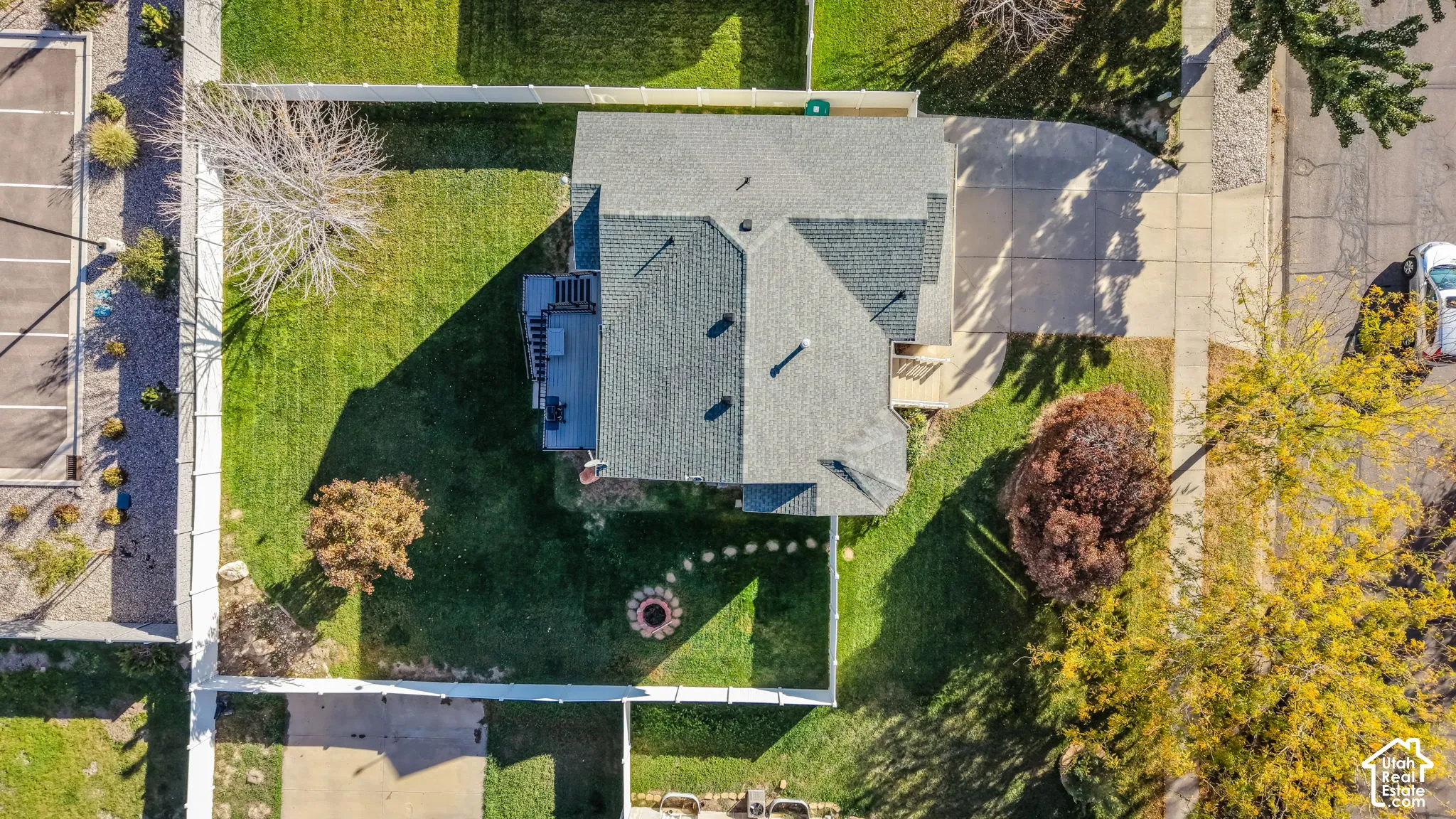 Aerial perspective featuring a shingled roof, concrete driveway, fenced yard, mature trees, and a stone-bordered fire pit