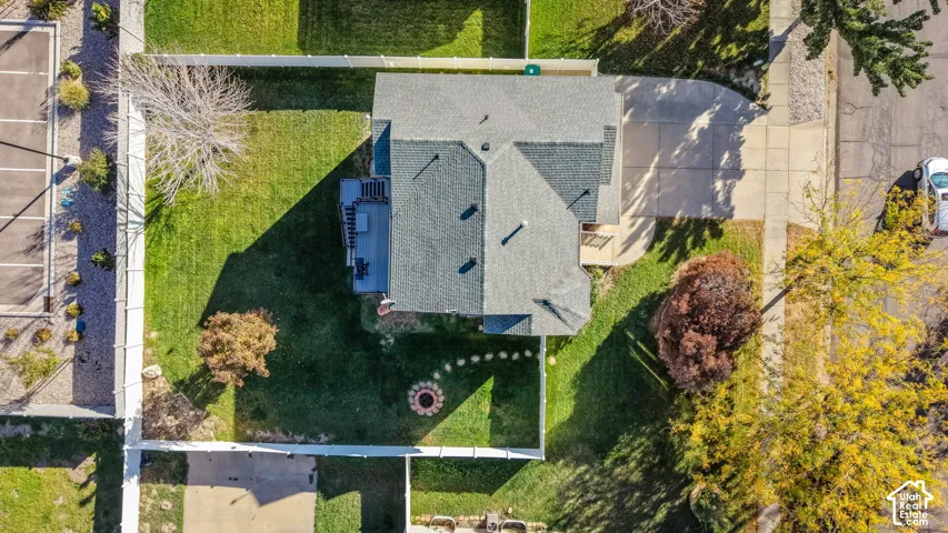 Aerial perspective featuring a shingled roof, concrete driveway, fenced yard, mature trees, and a stone-bordered fire pit