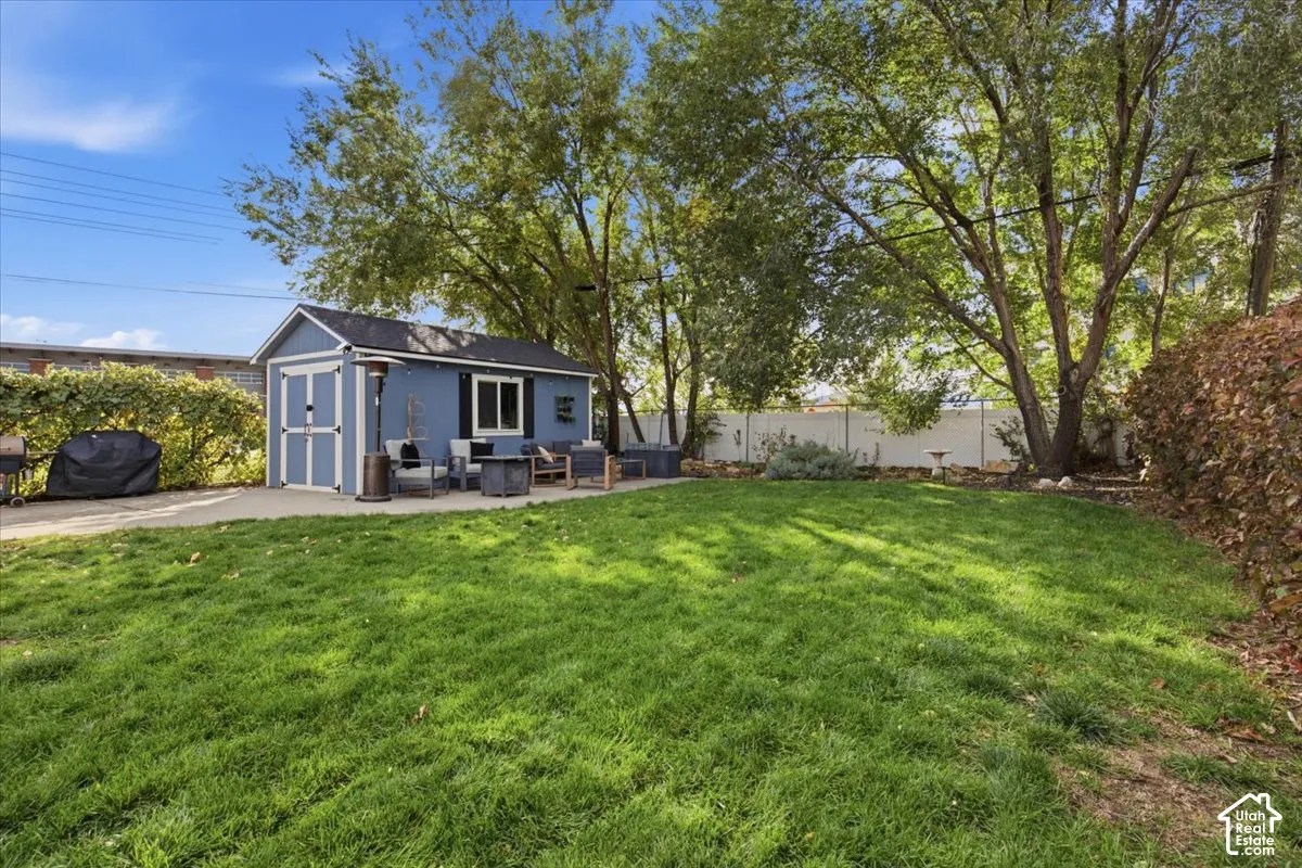 Backyard featuring a concrete patio, detached storage structure with white trim, mature trees, and perimeter fencing