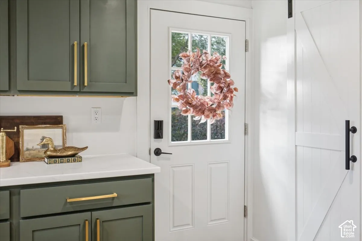 Olive-green cabinetry with gold-tone hardware, white countertop, white exterior door with glass insert, and white sliding barn door featuring a matte black pull handle