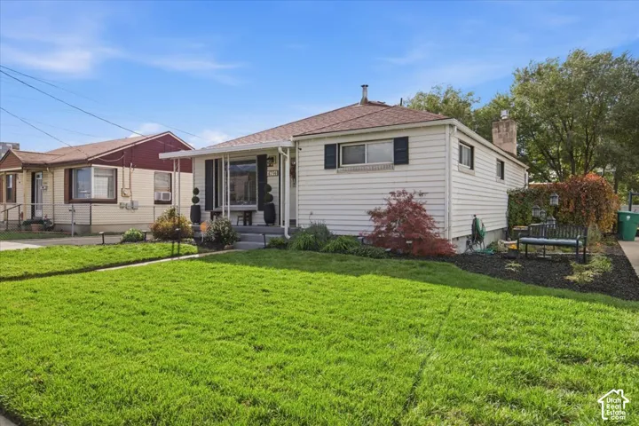 Single-story residence featuring light-colored horizontal siding, a covered front entryway, black window shutters, a chimney, and a landscaped front lawn