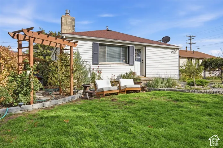 Exterior featuring white siding, a stone chimney, wooden garden trellis, and a raised stone landscape border