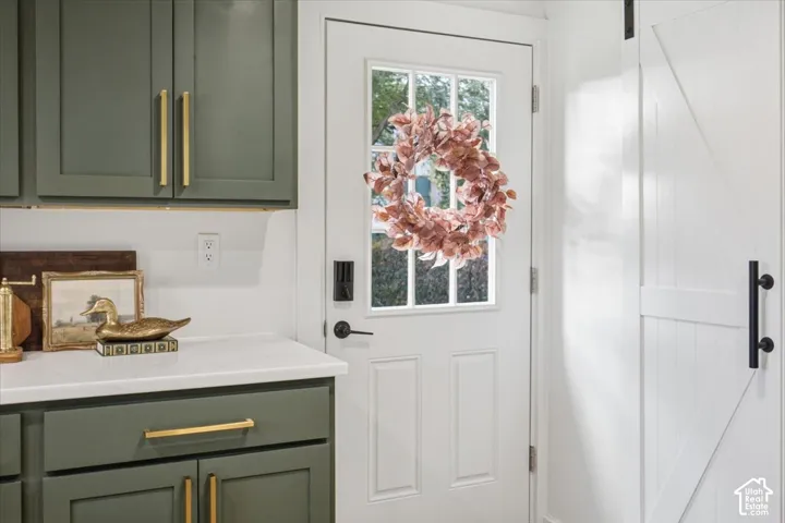 Olive-green cabinetry with gold-tone hardware, white countertop, white exterior door with glass insert, and white sliding barn door featuring a matte black pull handle
