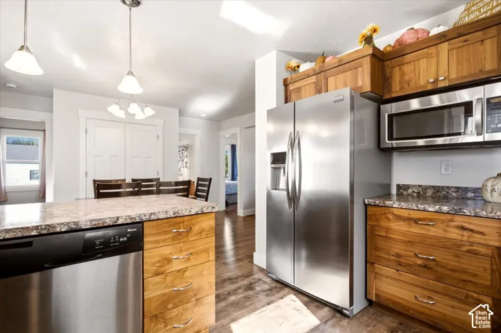 Kitchen featuring natural wood cabinetry, stone-patterned countertops, stainless steel appliances, wood-finish flooring, and multi-bulb pendant lighting