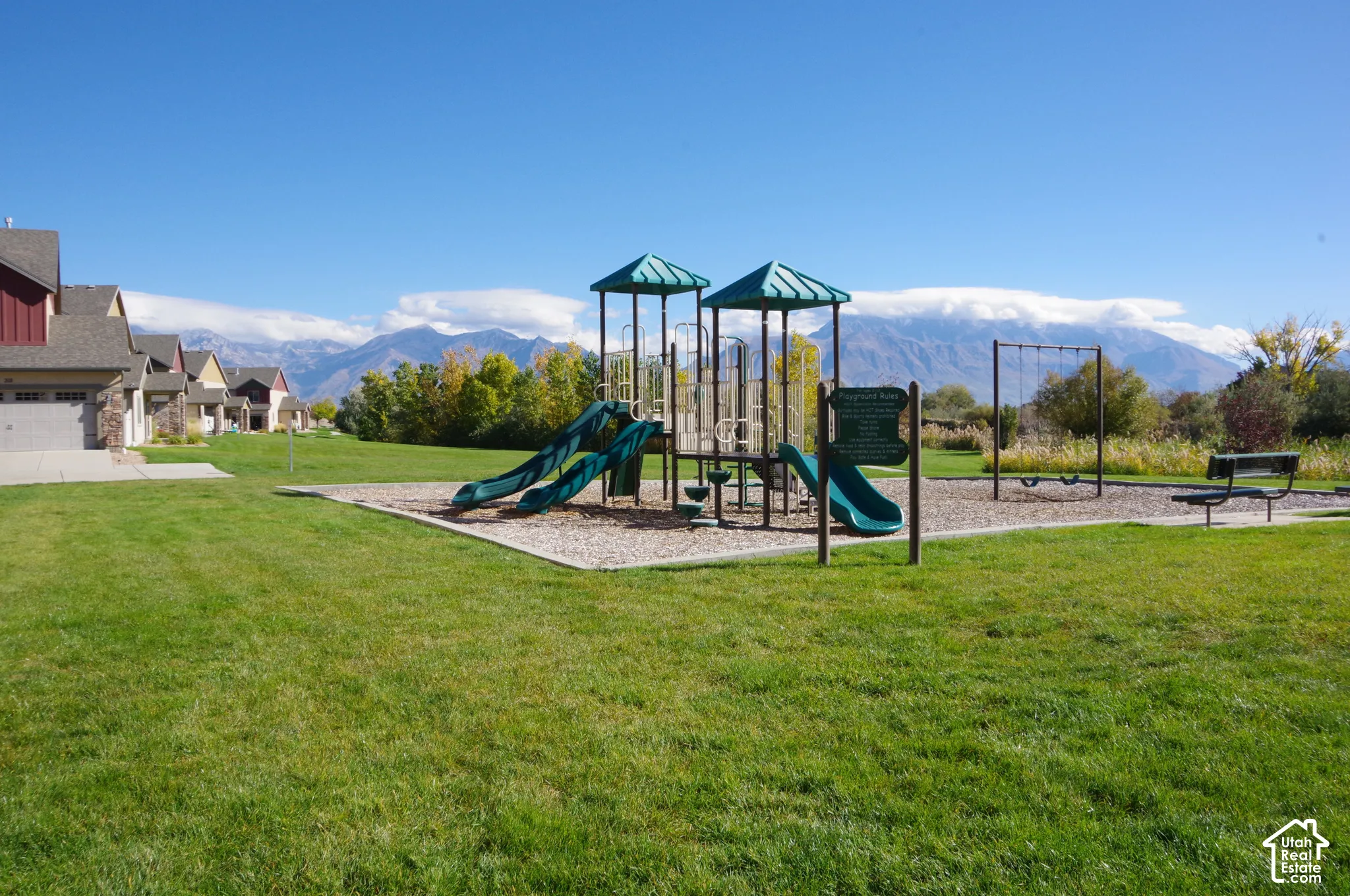 Outdoor recreation area featuring a playground structure with dual slides and a swing set, expansive grass lawn, and a mountain backdrop