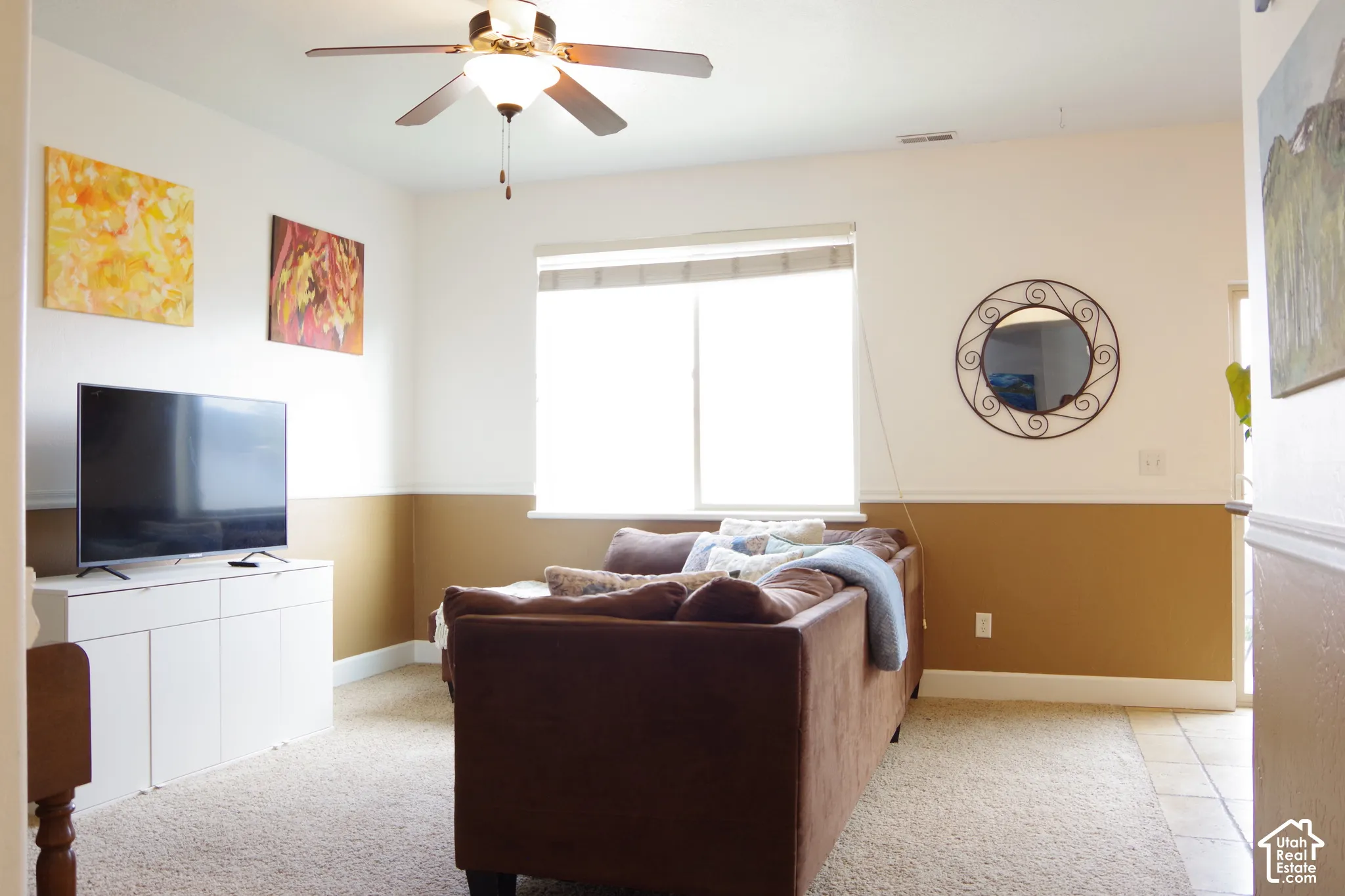 Neutral-toned interior featuring dual-tone wall paint, chair rail molding, wall-to-wall carpeting, a center ceiling fan with integrated lighting, and a window with blinds
