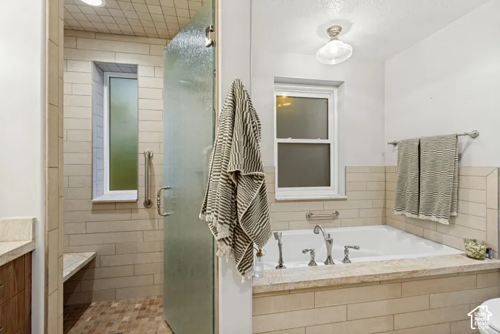 Bathroom featuring a built-in soaking tub with stone surround, a walk-in shower with textured glass door, horizontal wall tiling, and metal hardware