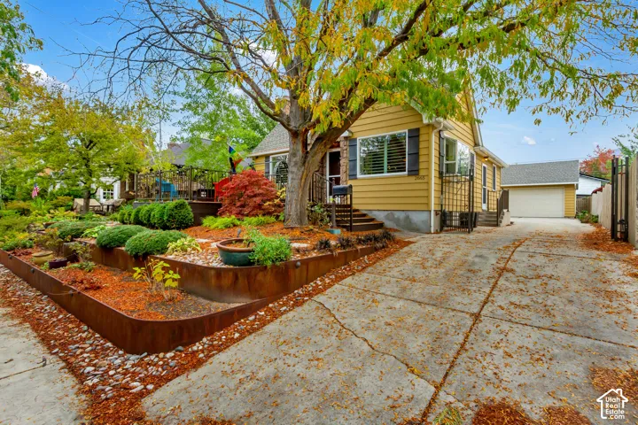 Yellow horizontal siding exterior featuring dark-toned shutters, a detached garage, tiered steel landscape edging, and a concrete driveway