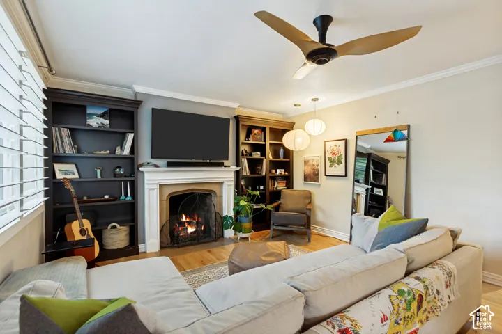 Fireplace with decorative mantle, built-in shelving, hardwood flooring, and three-blade ceiling fan
