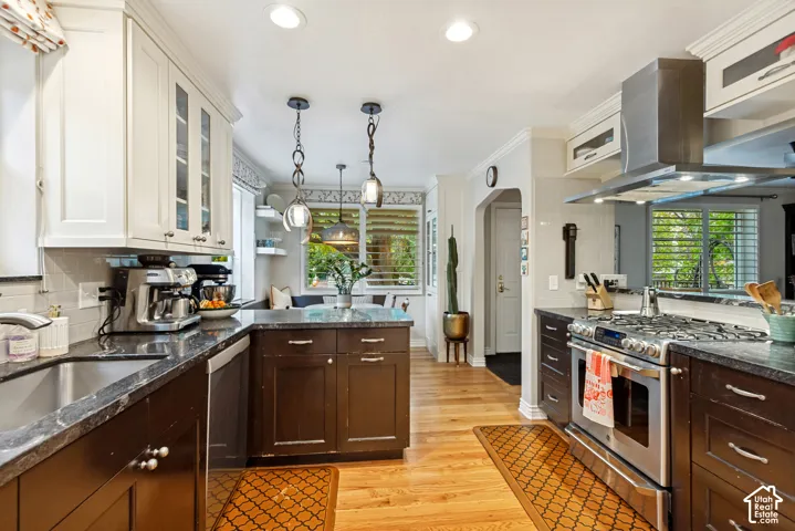 Kitchen featuring custom dark-toned lower cabinetry, white upper cabinets with glass-front accents, stainless steel range and hood, stone-surface countertops, and wood-finish flooring