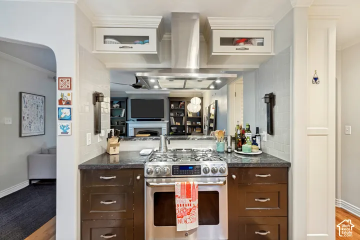 Stainless steel gas range and chimney-style range hood, dark stone-finish countertops, subway tile backsplash, and white shaker-style cabinetry with glass-paneled upper storage