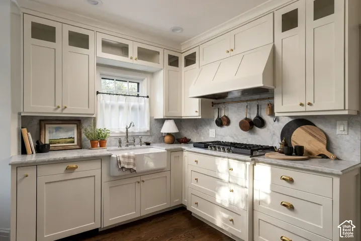 Kitchen featuring shaker-style cabinetry with glass-front uppers, apron-front farmhouse sink, stone backsplash and countertops, gas cooktop with custom range hood, and brass-finish hardware