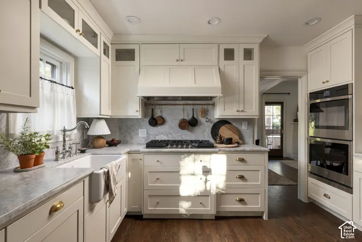 Custom cabinetry featuring glass-front upper displays, farmhouse sink with bridge-style faucet, stone countertops, integrated range hood, and double wall ovens