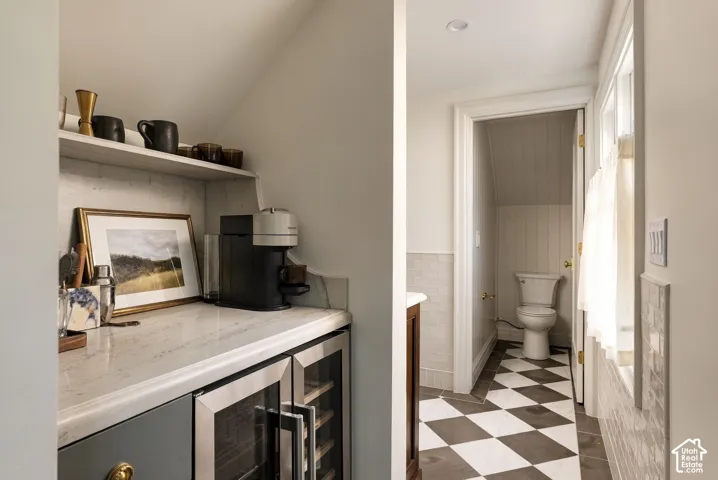 Wet bar featuring quartz countertops, built-in under-counter beverage refrigeration, and recessed shelving, accompanied by a bathroom with checkered tile flooring and wainscoting