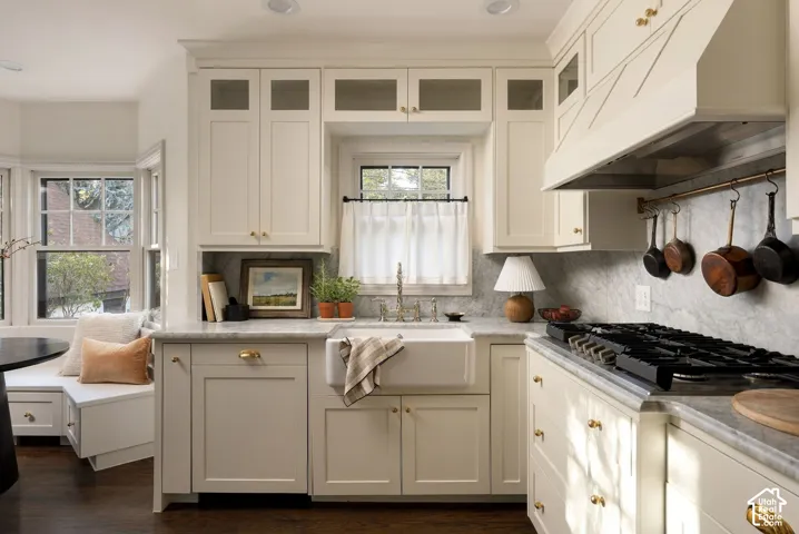 Kitchen featuring custom cabinetry with glass-front uppers, an apron-front farmhouse sink, stone countertops, integrated range hood, and built-in window seating