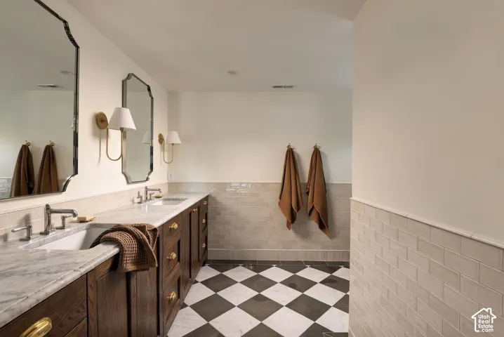 Double vanity featuring natural stone countertops, under-mount sinks, custom wood cabinetry with brass hardware, subway tile wainscoting, and checkered tile flooring