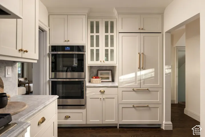 Custom off-white cabinetry featuring glass-front display doors and brass-finish hardware, stainless steel double wall ovens, panel-ready refrigerator, stone-composite countertops, and dark hardwood flooring
