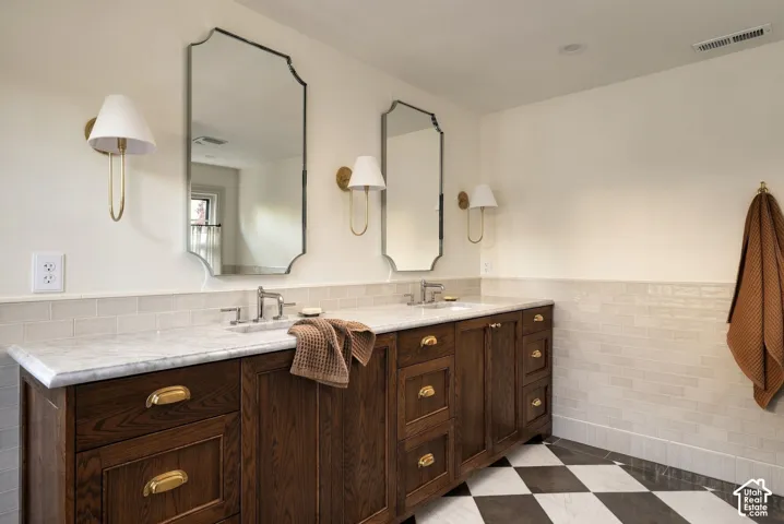 Double vanity featuring dark wood cabinetry, light-toned stone countertops, polished brass hardware, decorative wall sconces, and custom-shaped mirrors