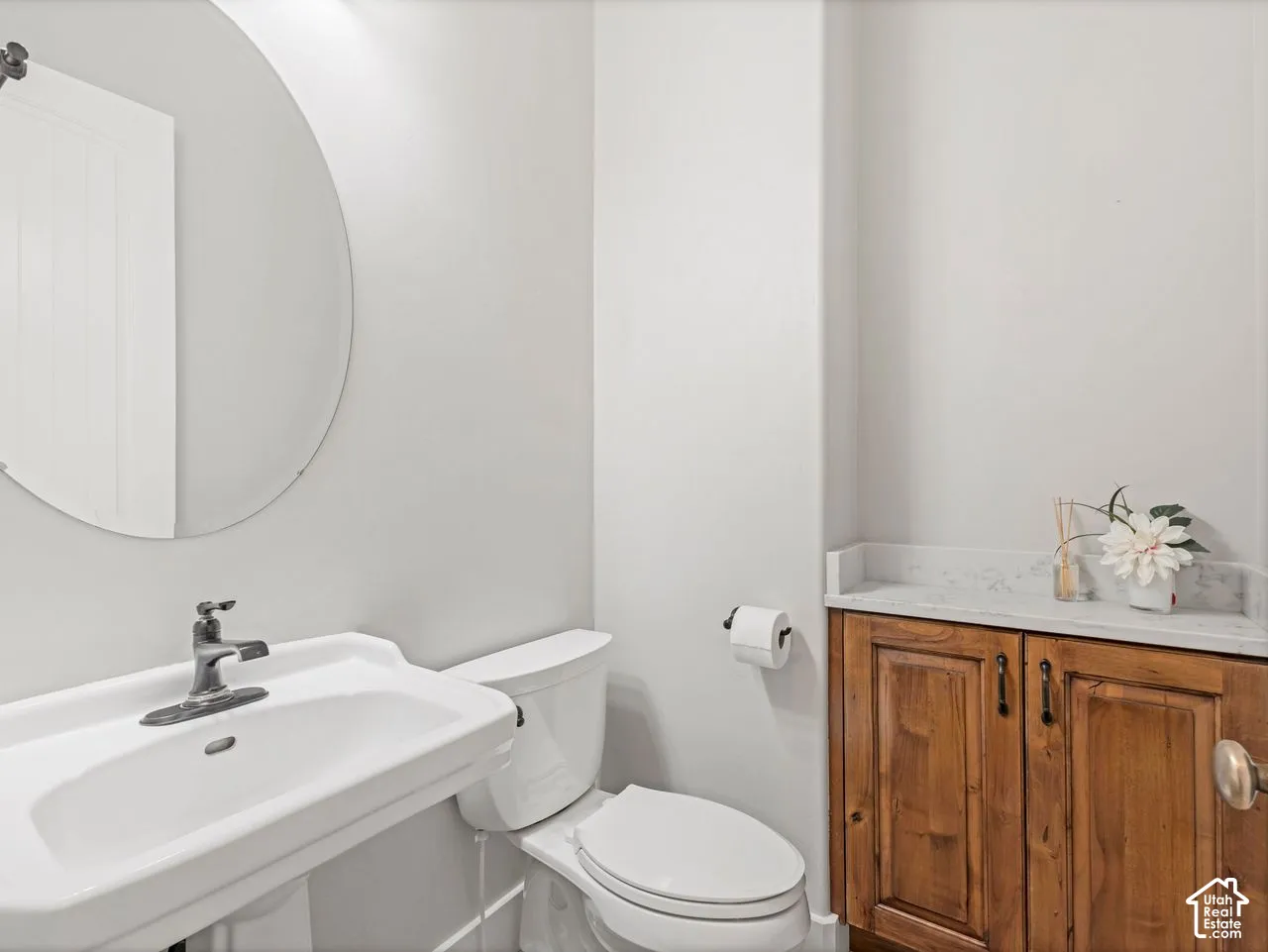 Pedestal sink with a metallic faucet, circular wall mirror, toilet, and wooden cabinetry topped with stone-finish surfacing