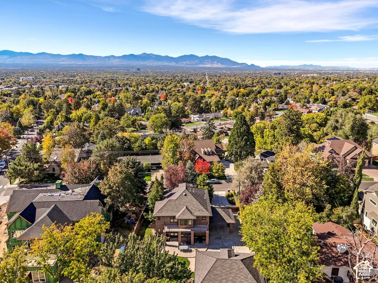 Aerial perspective featuring mountain range backdrop, established neighborhood with mature tree canopy, and brick-exterior residence with paved hardscaping