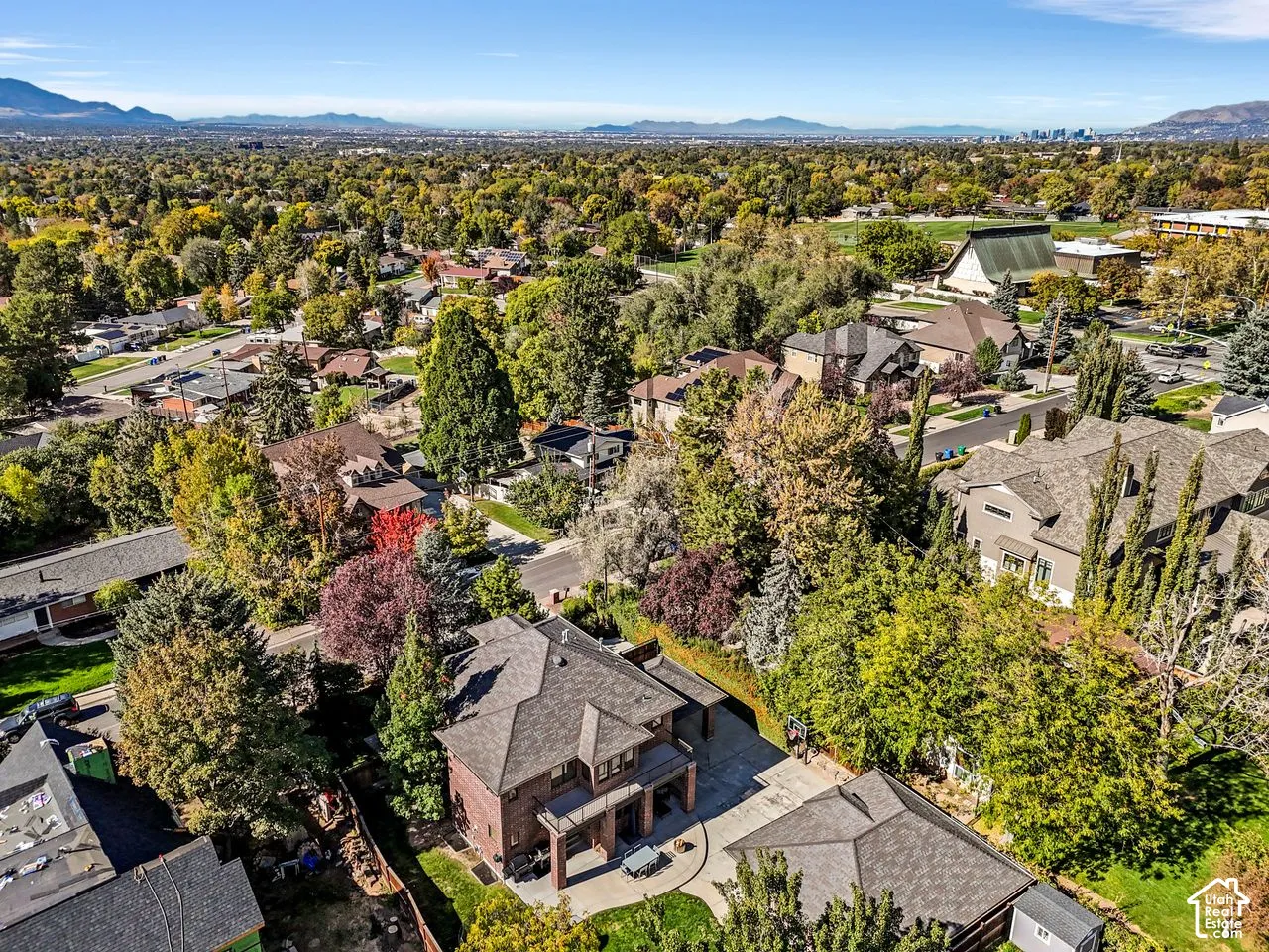 Aerial perspective featuring a multi-story brick residence with an expansive concrete patio and mature trees, set against a backdrop of distant mountains