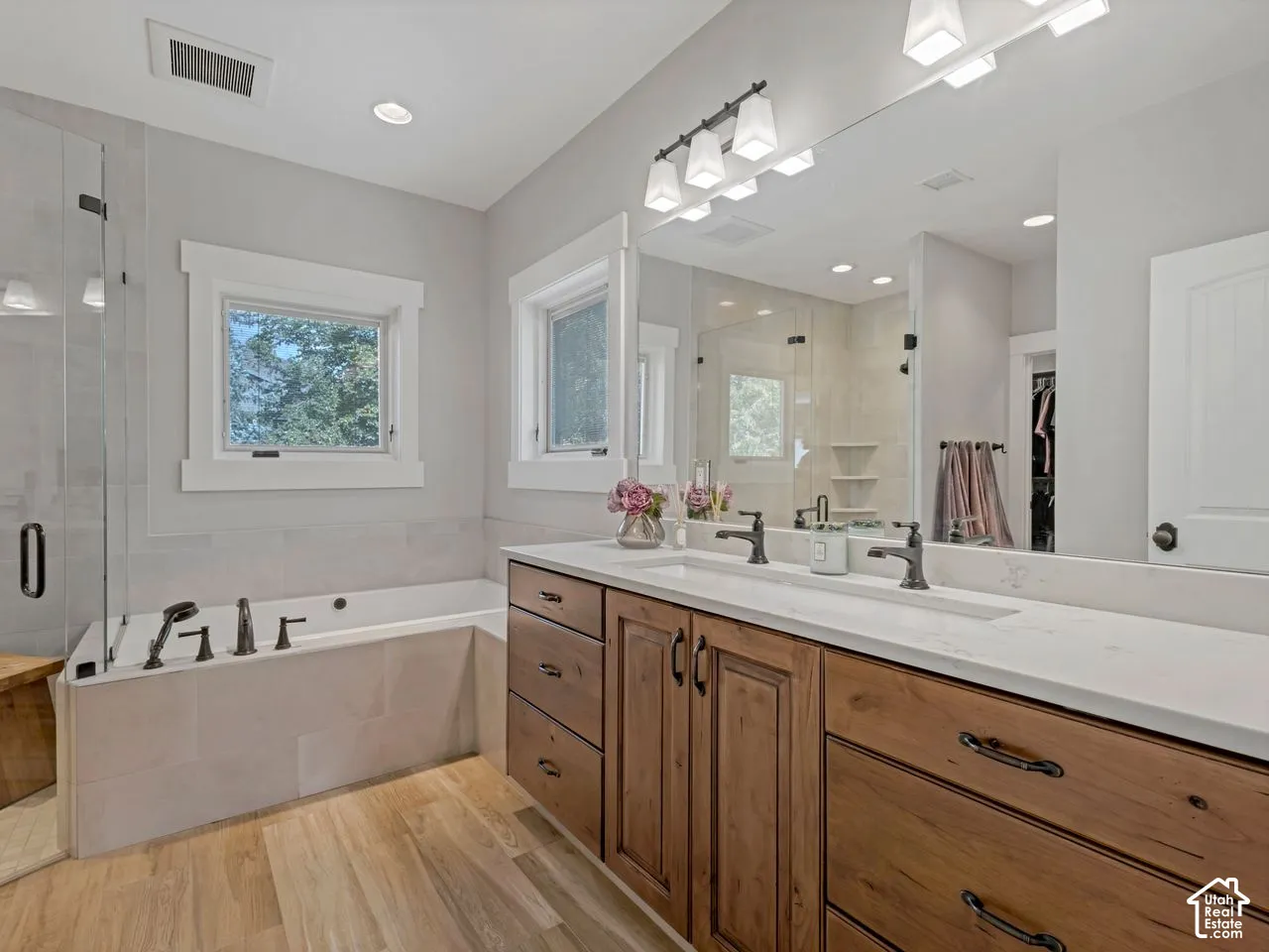 Dual-sink vanity with light-toned countertop, wood-finish cabinetry, frameless glass shower, built-in soaking tub, and wide-plank wood-finish flooring