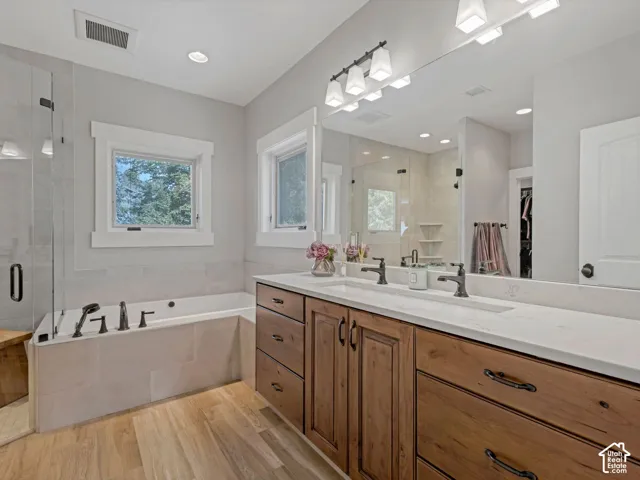 Dual-sink vanity with light-toned countertop, wood-finish cabinetry, frameless glass shower, built-in soaking tub, and wide-plank wood-finish flooring