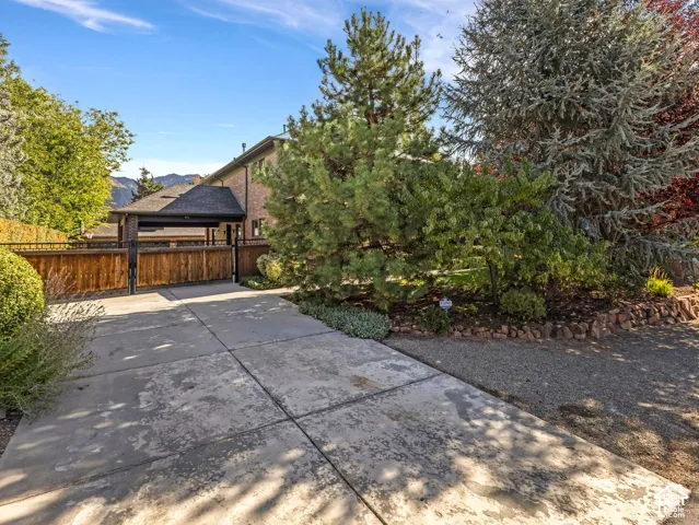 Concrete driveway leading to a covered parking area with a wood-paneled privacy gate, complemented by mature evergreen landscaping and a stone-bordered garden bed