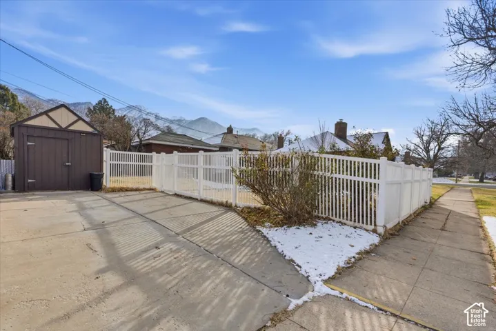Concrete driveway with a detached storage shed, enclosed by white vinyl fencing and featuring a mountain backdrop