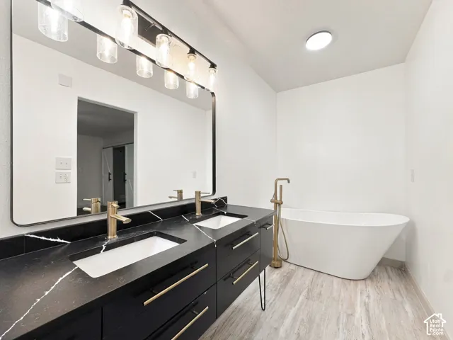 Dual-sink vanity with dark stone-finish countertops, brass-toned fixtures, a large rectangular mirror, and a freestanding soaking tub with a floor-mounted brass-toned faucet, complemented by wood-finish flooring