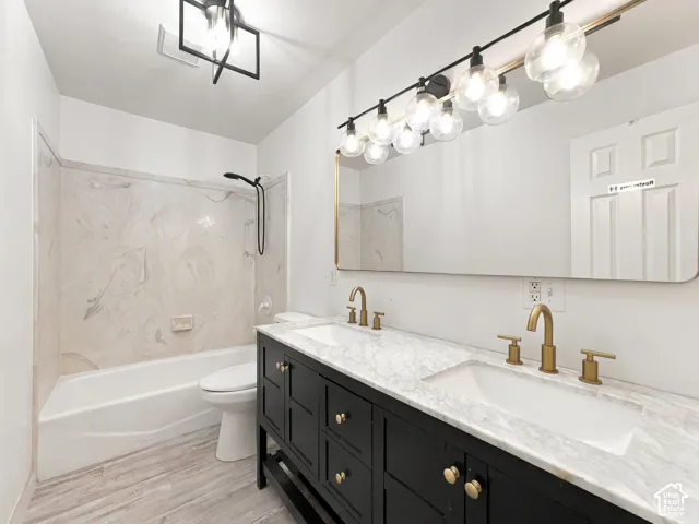 Double vanity featuring black cabinetry, stone-patterned countertop, gold-tone hardware, multi-bulb light fixture, and a patterned tub surround