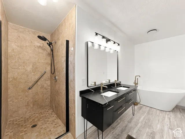 Walk-in shower with tile surround, black dual-vanity cabinet with stone countertop, freestanding soaking tub, wood-finish flooring, and minimalist vanity lighting