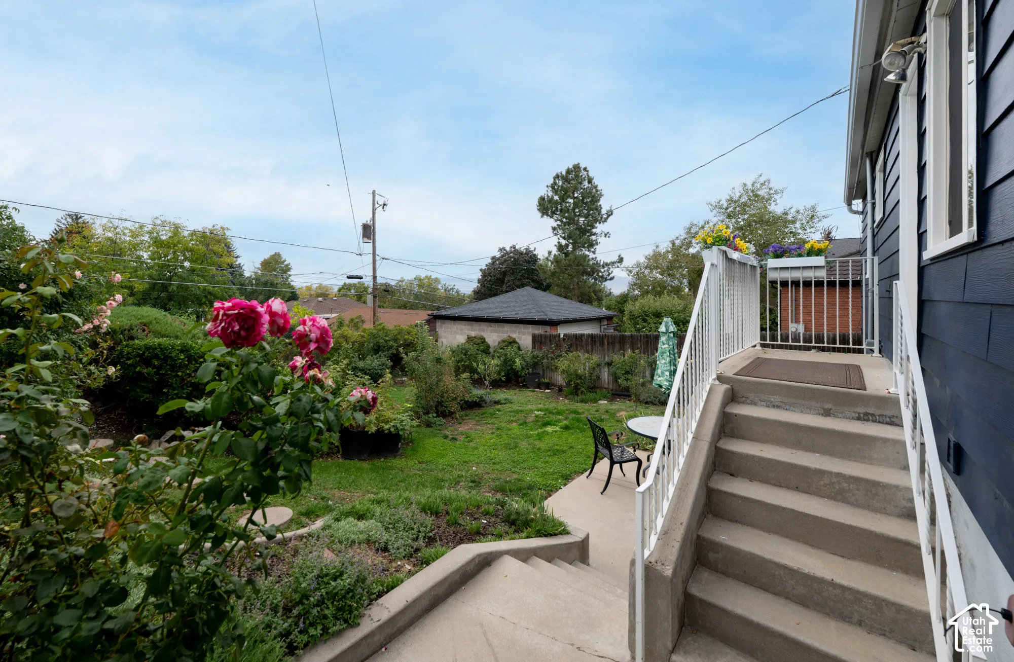 Rear exterior featuring concrete steps with metal railings, dark blue horizontal siding, and a landscaped backyard with mature plantings and a lawn