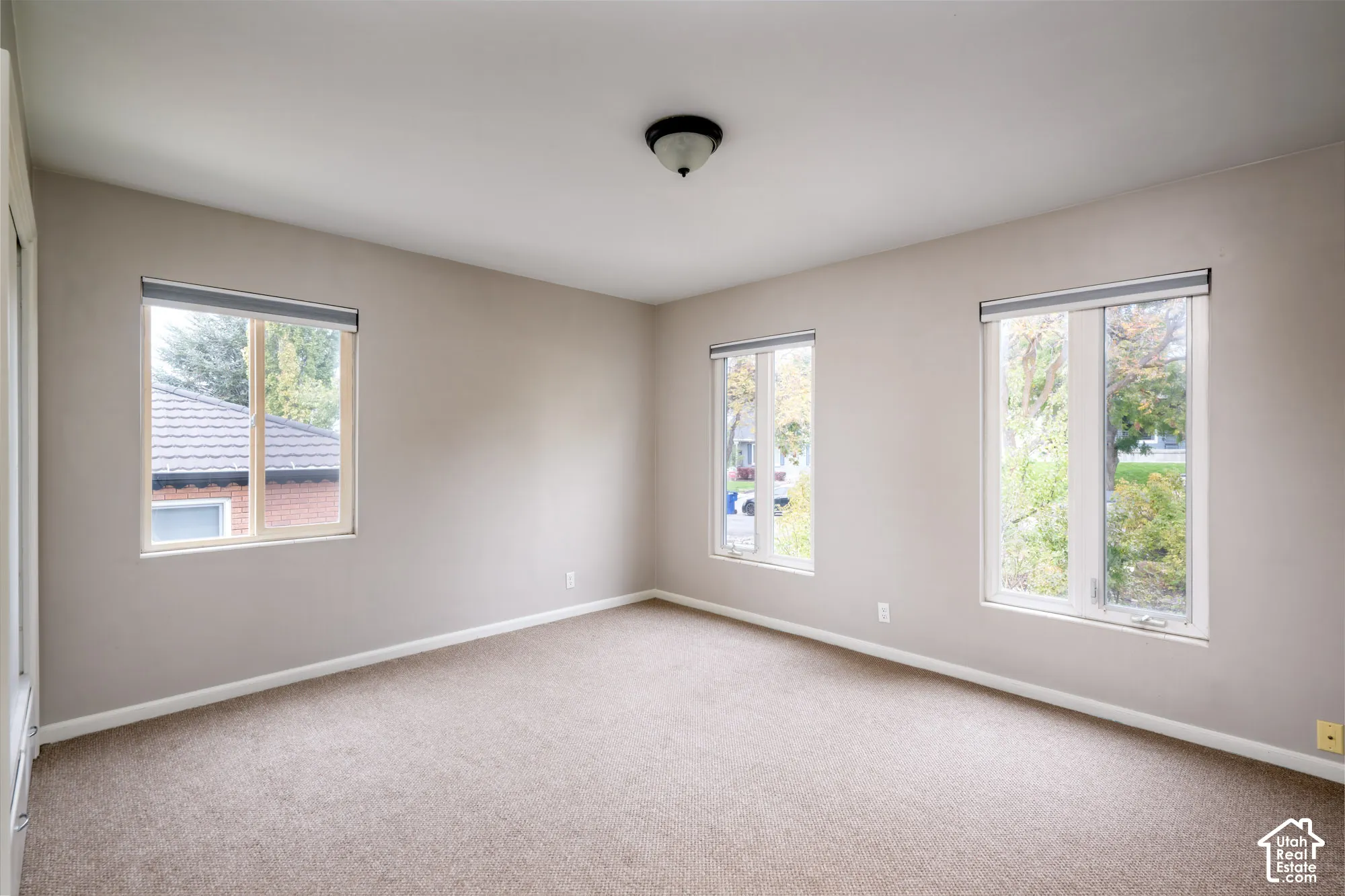 Neutral-toned interior featuring wall-to-wall carpeting, three windows with roller shades, and a central flush-mount ceiling light fixture