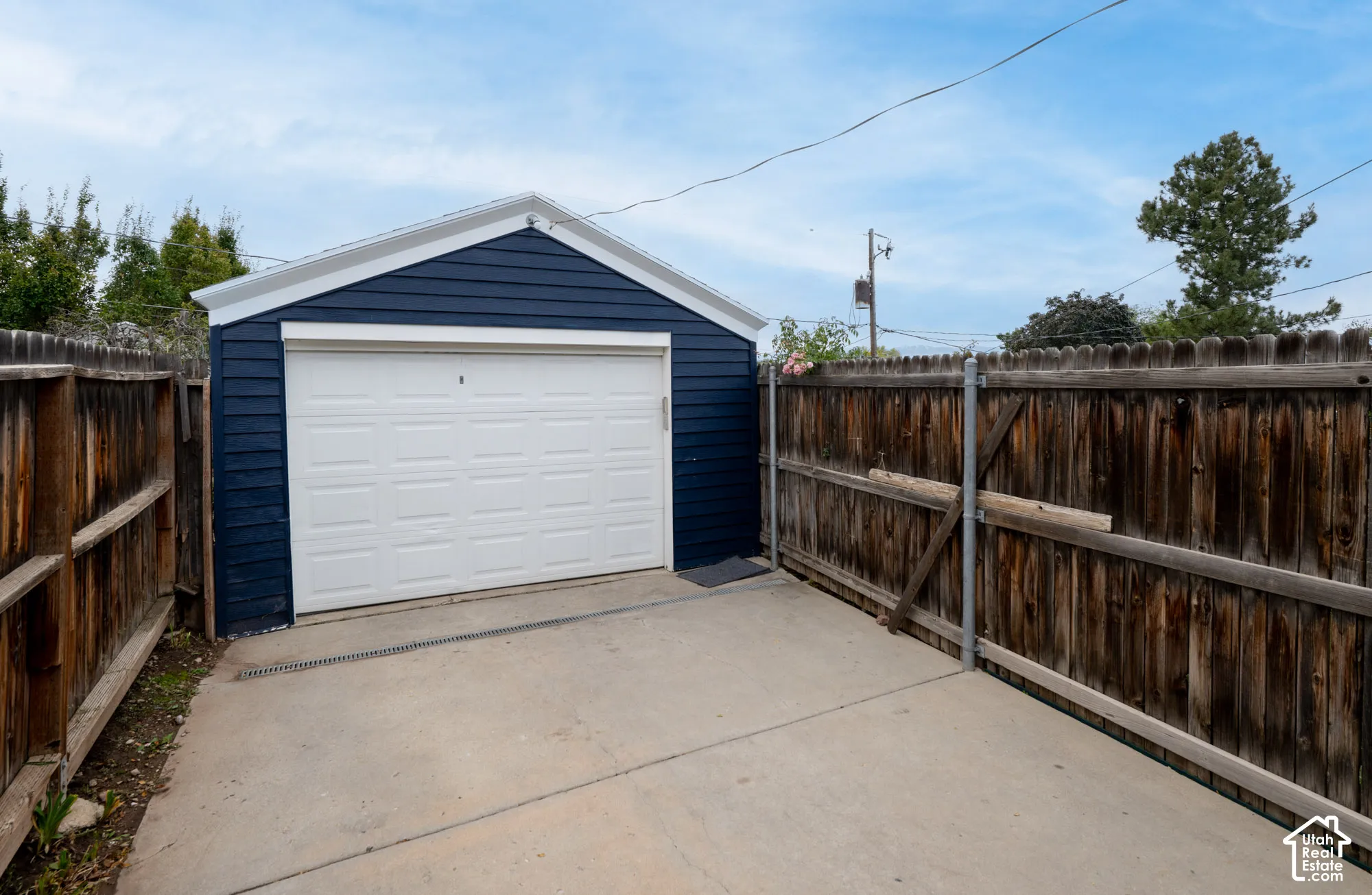 Detached garage with navy blue horizontal siding, white sectional door, and concrete apron enclosed by perimeter wood fencing